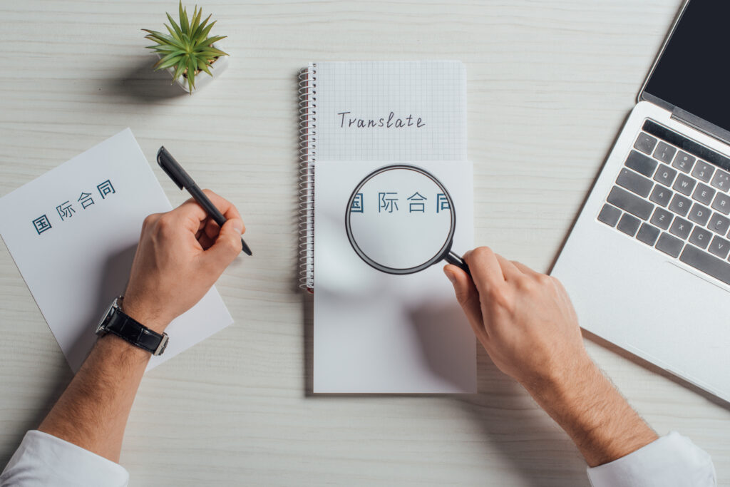 Manos de una persona sosteniendo una lupa y un bolígrafo para hacer traducción japonés junto a una laptop