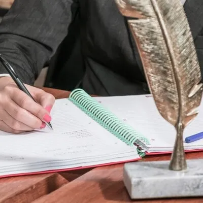 Fotografía de una persona escribiendo en una libreta y enfrente una pluma que es ícono de una agencia de traducción.