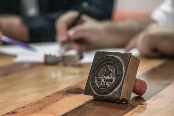 Fotografía de un sello sobre una mesa de madera y personas escribiendo al fondo en una agencia de traducción.