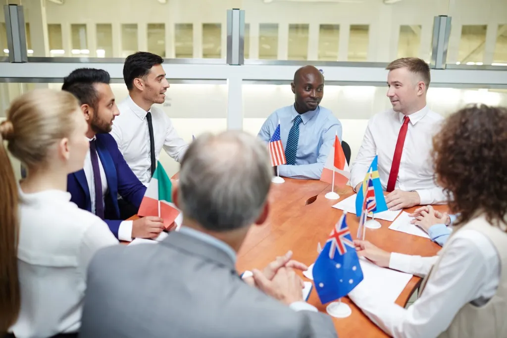 Comunicación institucional: fotografía de grupo de personas sentadas alrededor de una mesa con banderas de distintos países.