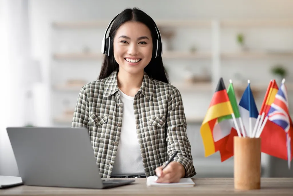 Traducción con IA: fotografía de una mujer utilizando audífonos frente a una computadora y a lado banderas de países.